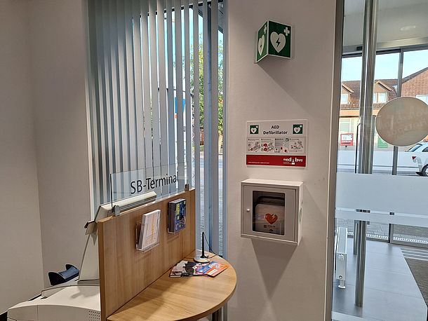 AED im Wandkasten an der Säule neben der Glastür (Ein-/Ausgang)